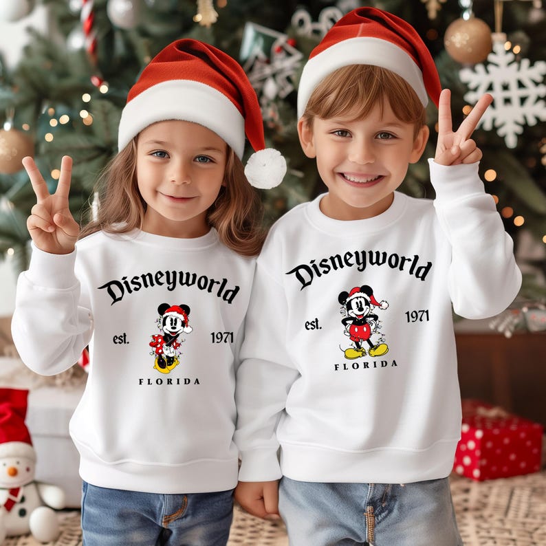 May include: Two children wearing white sweatshirts with a Mickey Mouse design and the text "Disneyworld est. 1971 Florida". The children are wearing red Santa hats and smiling.
