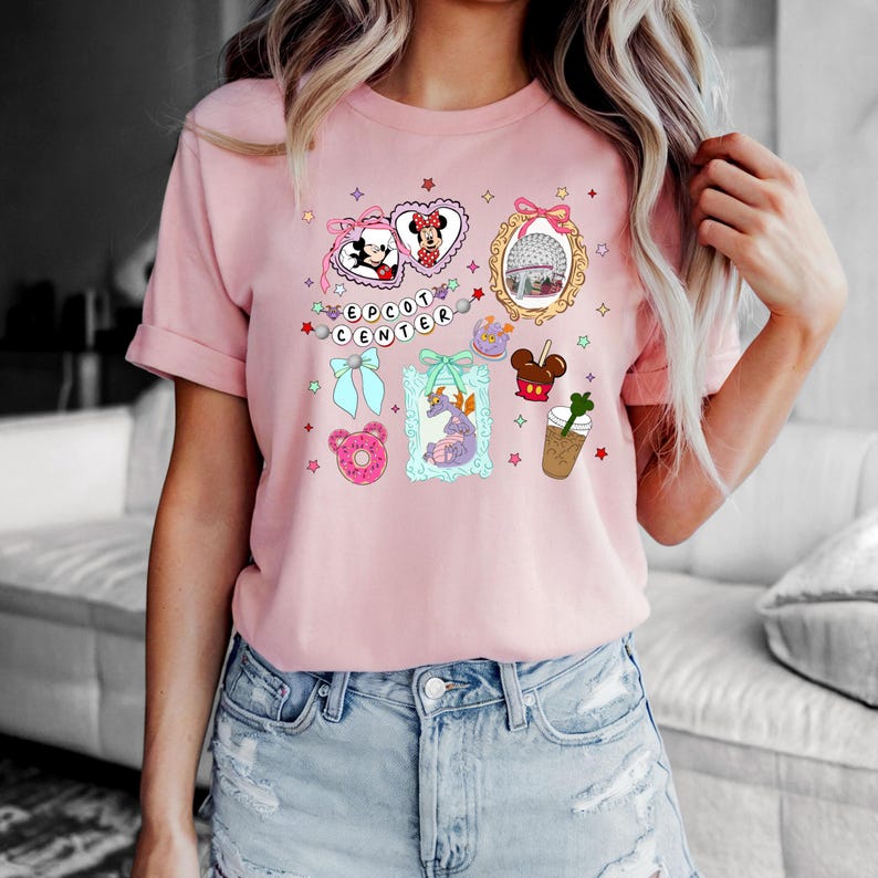 May include: Pink t-shirt with a graphic design featuring various Disney characters and icons, including Mickey Mouse, Minnie Mouse, a castle, and a donut. The text "EPCOT CENTER" is also included in the design.
