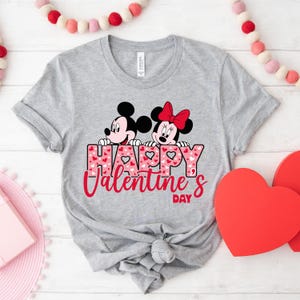 May include: A grey t-shirt featuring a graphic of Mickey Mouse and Minnie Mouse peeking over the words "Happy Valentine's Day" with a pink and white heart pattern.