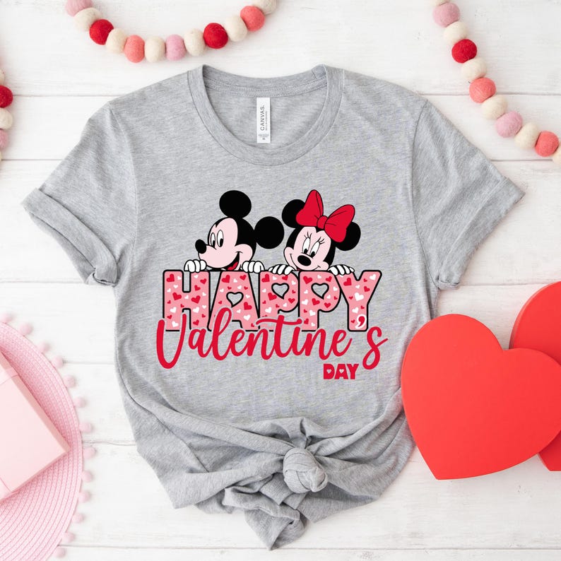 May include: A grey t-shirt featuring a graphic of Mickey Mouse and Minnie Mouse peeking over the words "Happy Valentine's Day" with a pink and white heart pattern.