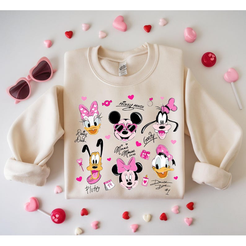 May include: Cream-colored sweatshirt featuring a collage of Disney character faces, including Mickey and Minnie Mouse, Goofy, Daisy and Donald Duck, and Pluto. Pink hearts and text accents complete the design. Heart-shaped sunglasses and candies surround the sweatshirt.