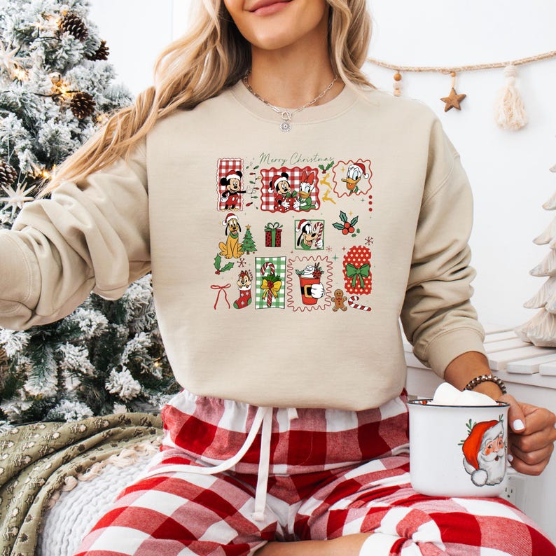 May include: Beige crewneck sweatshirt featuring a colorful Christmas-themed graphic with Disney characters and the text "Merry Christmas." The graphic includes Mickey Mouse, Minnie Mouse, Donald Duck, and other festive elements. The person is wearing red and white plaid pajama pants.
