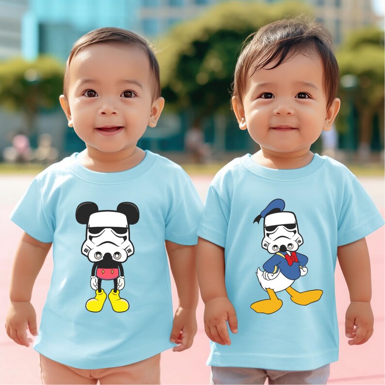 May include: Two light blue baby t-shirts. One features a Stormtrooper helmet on Mickey Mouse, the other on Donald Duck. The cartoon characters are in black, white, red, and yellow. The shirts are short-sleeved.