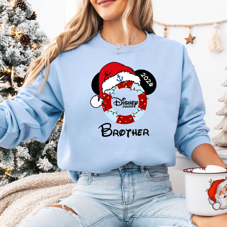 K&ouml;nnte beinhalten: Hellblaues Sweatshirt mit festlichem Disney Cruise-Design. Das Design umfasst einen Weihnachtsmannhut, Mickey Mouse-Ohren, eine Rettungsboje mit der Aufschrift "Disney Cruise" und das Wort "Brother". Das Jahr "2026" ist ebenfalls enthalten.