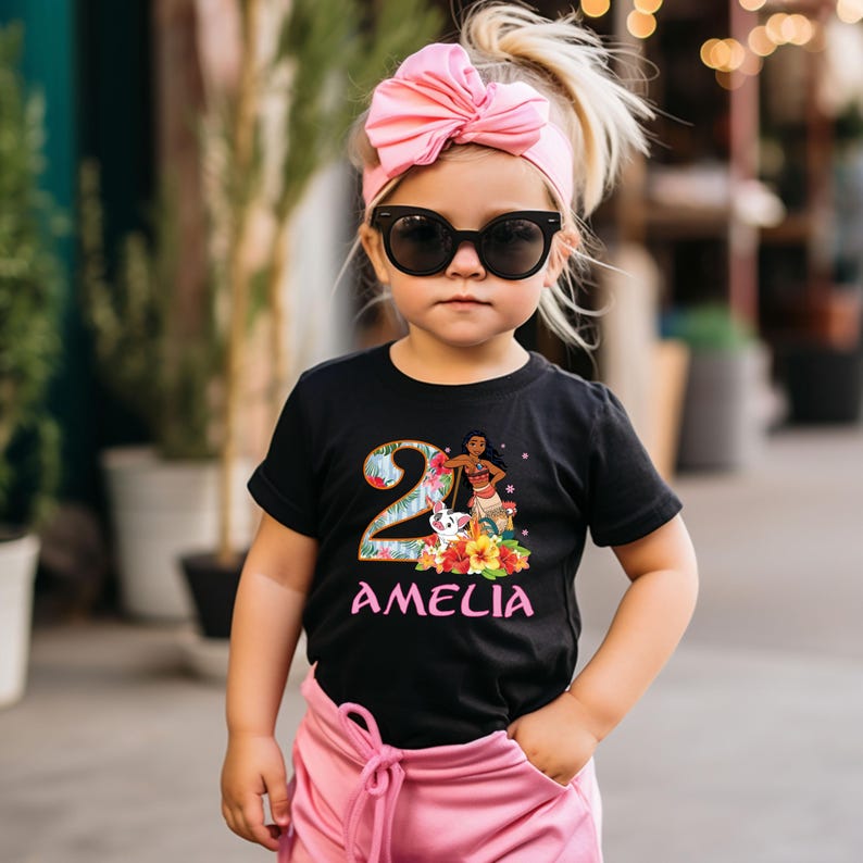 May include: A black t-shirt with a colorful graphic featuring the number 2, a cartoon character, and the name "AMELIA" in pink. The shirt is worn by a young child wearing sunglasses and a pink bow headband. The child is also wearing pink pants.