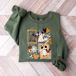 Puede incluir: Sudadera verde con un dise&ntilde;o tem&aacute;tico de Halloween. El dise&ntilde;o incluye fantasmas de dibujos animados, calabazas, un castillo y una taza de caf&eacute;. Tambi&eacute;n son visibles las palabras "Minnie Magic". La sudadera est&aacute; doblada sobre una superficie de madera.