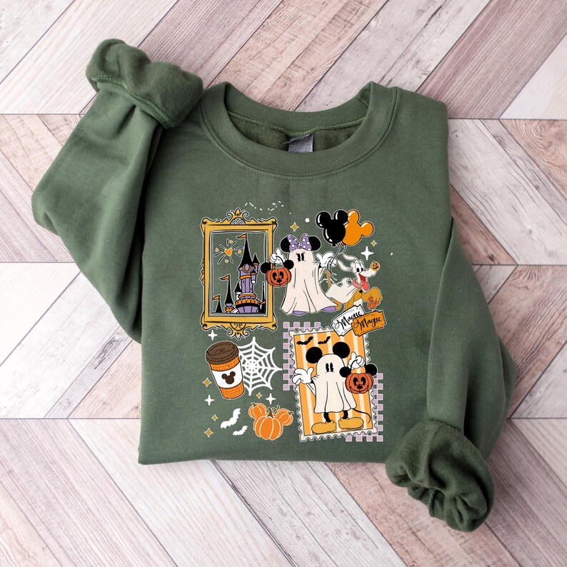 Puede incluir: Sudadera verde con un dise&ntilde;o tem&aacute;tico de Halloween. El dise&ntilde;o incluye fantasmas de dibujos animados, calabazas, un castillo y una taza de caf&eacute;. Tambi&eacute;n son visibles las palabras "Minnie Magic". La sudadera est&aacute; doblada sobre una superficie de madera.