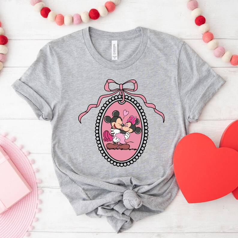 K&ouml;nnte beinhalten: Ein graues T-Shirt mit einem rosa und schwarzen Design von Mickey Mouse und Minnie Mouse, die sich in einem herzf&ouml;rmigen Rahmen k&uuml;ssen. Der Rahmen ist mit einer rosa Schleife und Perlen verziert.