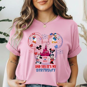May include: Pink t-shirt featuring a Mickey Mouse design with Daisy Duck, Donald Duck, Mickey Mouse, and Minnie Mouse. The design includes a castle, hearts, fireworks, and the text "4th of July AND YES IT'S MY BIRTHDAY."