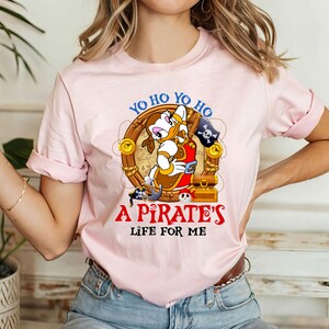 K&ouml;nnte beinhalten: Pinkes T-Shirt mit einem Cartoon-Enten-Piraten-Charakter, der ein rot-goldenes Outfit tr&auml;gt. Der Text "Yo ho yo ho A Pirate's Life For Me" ist auf das Shirt gedruckt.