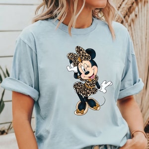 Op de afbeelding: Een lichtblauw T-shirt met een afbeelding van Minnie Mouse die een luipaardprintjurk en een strik draagt.