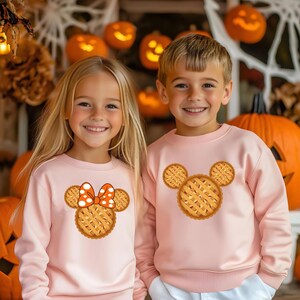 K&ouml;nnte beinhalten: Zwei Kinder in passenden hellrosa Sweatshirts. Jedes Sweatshirt hat ein Kuchen-Design in Form eines Mickey Mouse-Kopfes. Eines hat eine orangefarbene und wei&szlig;e gepunktete Schleife. K&uuml;rbisse und Halloween-Dekorationen im Hintergrund.