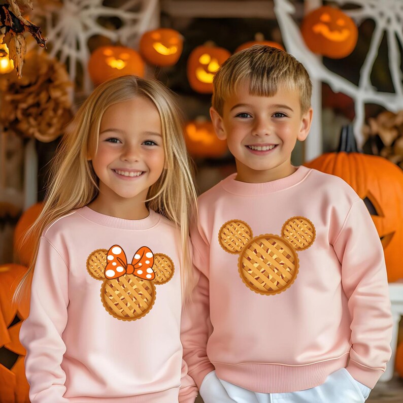 K&ouml;nnte beinhalten: Zwei Kinder in passenden hellrosa Sweatshirts. Jedes Sweatshirt hat ein Kuchen-Design in Form eines Mickey Mouse-Kopfes. Eines hat eine orangefarbene und wei&szlig;e gepunktete Schleife. K&uuml;rbisse und Halloween-Dekorationen im Hintergrund.