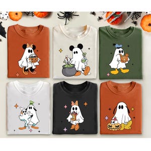 May include: Six long-sleeved t-shirts in various colors, each featuring a cartoon ghost character with Mickey Mouse ears. The ghosts are holding pumpkins, cauldrons, and potion bottles. Halloween-themed apparel.