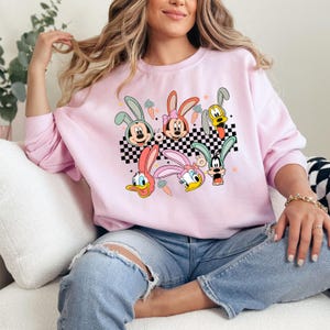 May include: Pink sweatshirt with a graphic print of cartoon characters with bunny ears. The characters are Mickey Mouse, Minnie Mouse, Donald Duck, and Goofy. The graphic is on a checkered background with carrots.