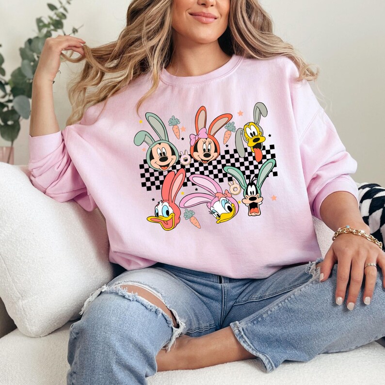 May include: Pink sweatshirt with a graphic print of cartoon characters with bunny ears. The characters are Mickey Mouse, Minnie Mouse, Donald Duck, and Goofy. The graphic is on a checkered background with carrots.