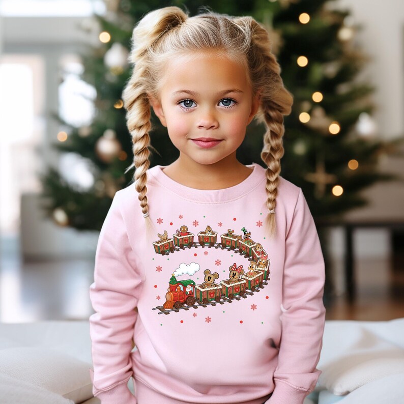 May include: A young girl wearing a light pink sweatshirt with a Christmas train design. The train features cartoon animals in each car, with snowflakes and stars scattered around the design. The background includes a Christmas tree.