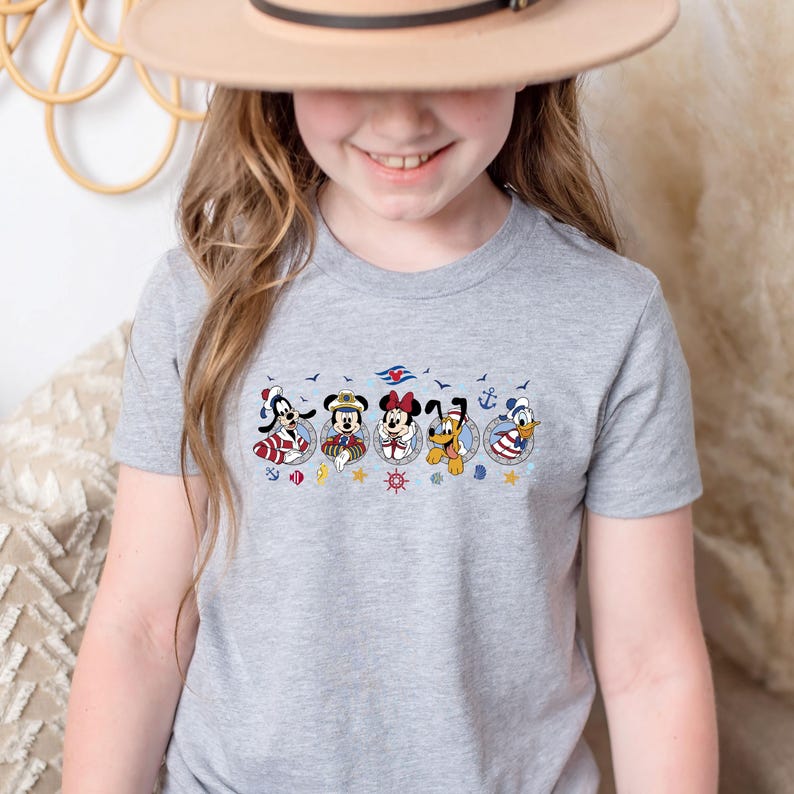 May include: A gray t-shirt featuring a colorful graphic of Disney characters in nautical-themed circles. The design includes Mickey Mouse, Minnie Mouse, Donald Duck, Goofy, and Pluto, with anchors and nautical elements.
