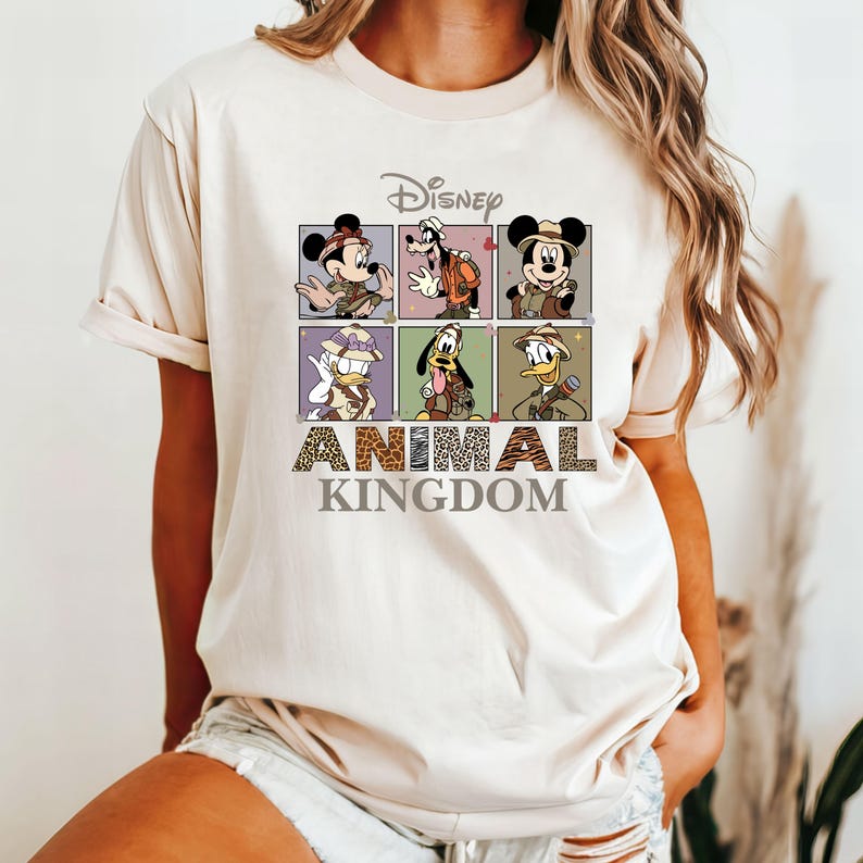 May include: A beige t-shirt with a graphic print of Mickey Mouse, Donald Duck, Goofy, and Pluto dressed as safari explorers. The text "Disney Animal Kingdom" is printed below the characters.
