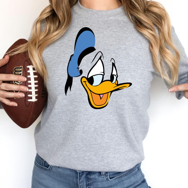 May include: A light gray sweatshirt featuring a cartoon image of Donald Duck's face. The design includes his blue sailor hat, yellow beak, and expressive eyes. The sweatshirt is paired with blue jeans.