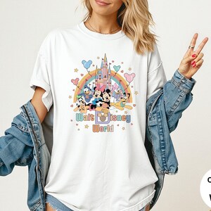 Puede incluir: Camiseta blanca con un gr&aacute;fico colorido de personajes de Disney, un arco&iacute;ris y un castillo. El texto "Walt Disney World" se muestra debajo. La camiseta tiene mangas cortas y un corte holgado.