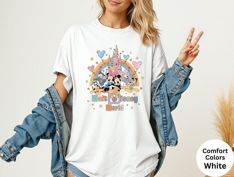 Puede incluir: Camiseta blanca con un gr&aacute;fico colorido de personajes de Disney, un arco&iacute;ris y un castillo. El texto "Walt Disney World" se muestra debajo. La camiseta tiene mangas cortas y un corte holgado.