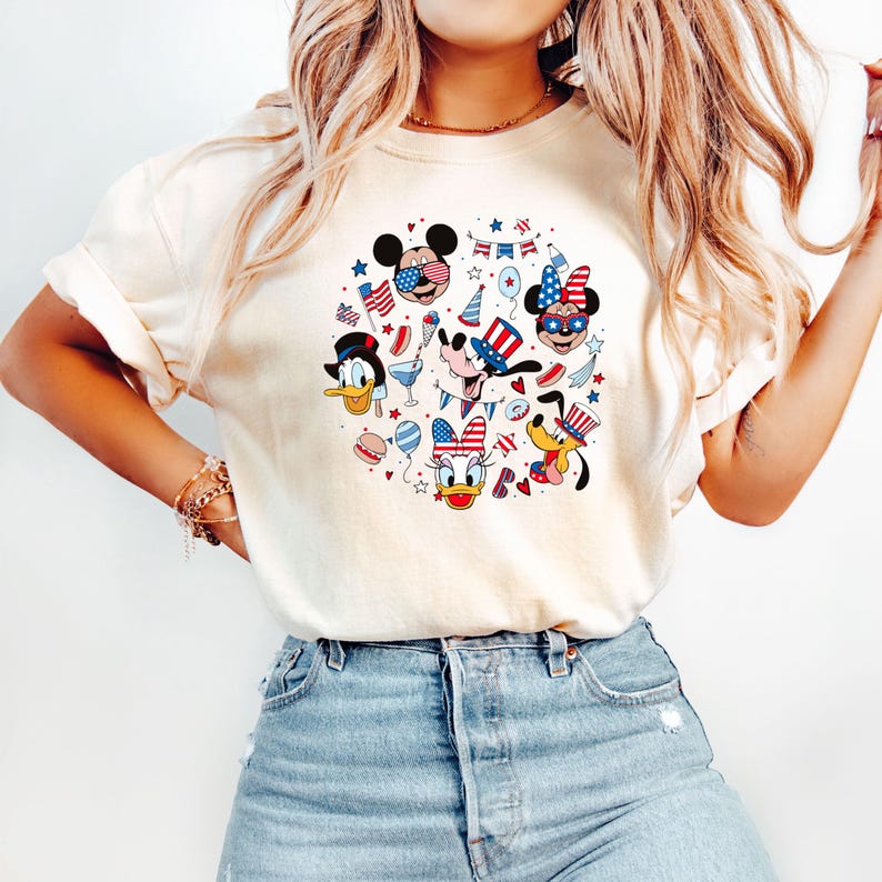 May include: Cream-colored t-shirt featuring a circular graphic of Disney characters in patriotic attire. Mickey Mouse, Minnie Mouse, Donald Duck, Goofy, and Pluto are depicted with American flag-themed accessories and festive decorations. The design includes stars, stripes, and celebratory elements.