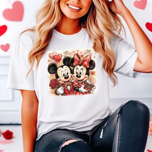 May include: White t-shirt featuring a colorful graphic of Mickey and Minnie Mouse in Valentine's Day attire. Mickey holds roses, and Minnie holds chocolates. Both are smiling and surrounded by hearts and balloons.