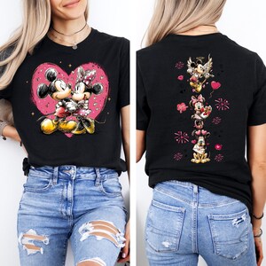 May include: Black t-shirt featuring Mickey and Minnie Mouse kissing inside a pink heart. The back of the shirt displays Mickey, Minnie, and Pluto with Valentine's Day graphics. The shirt is worn by a person.
