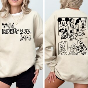 May include: A beige sweatshirt with a black and white graphic print of Mickey Mouse and friends. The front of the sweatshirt says "Mickey & Co.". The back of the sweatshirt has a larger graphic print of Mickey Mouse, Minnie Mouse, Donald Duck, and Goofy. The text on the back says "Mickey & Co. Est. 1928."
