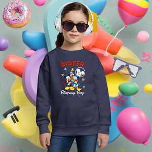 May include: A navy blue sweatshirt with the word "SISTER" in orange script and a graphic of Mickey Mouse, Donald Duck, and Goofy. The text "Birthday Boy" is below the graphic. The sweatshirt is worn by a person wearing sunglasses and headphones.
