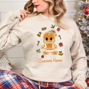 May include: A cream-colored sweatshirt with a gingerbread man wearing a Santa hat and a face mask. The gingerbread man is surrounded by Christmas-themed decorations, including candy canes, bells, and holly. The text "Custom Name" is printed below the gingerbread man.