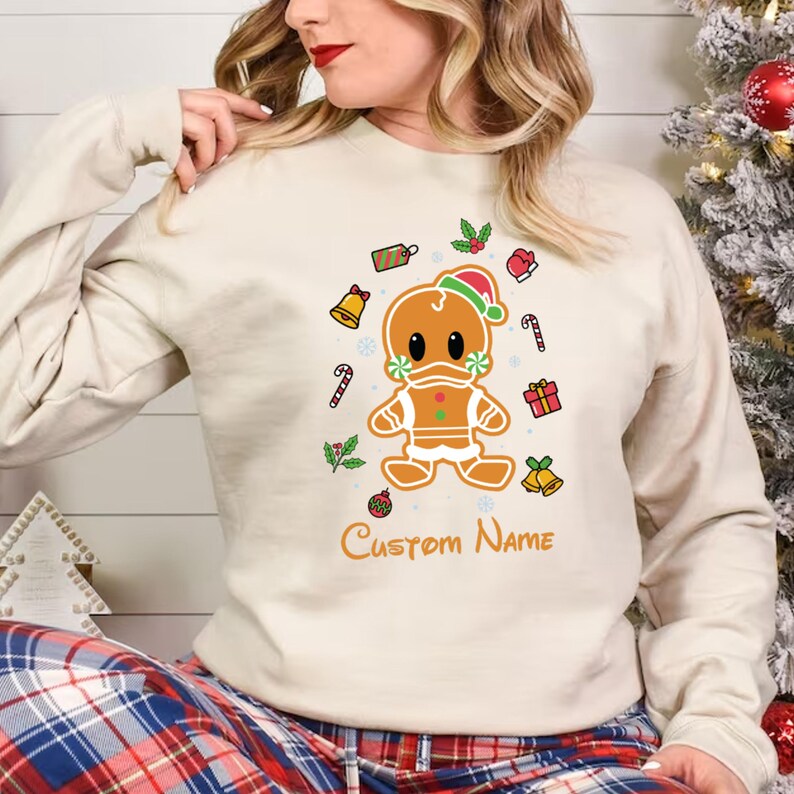 May include: A cream-colored sweatshirt with a gingerbread man wearing a Santa hat and a face mask. The gingerbread man is surrounded by Christmas-themed decorations, including candy canes, bells, and holly. The text "Custom Name" is printed below the gingerbread man.