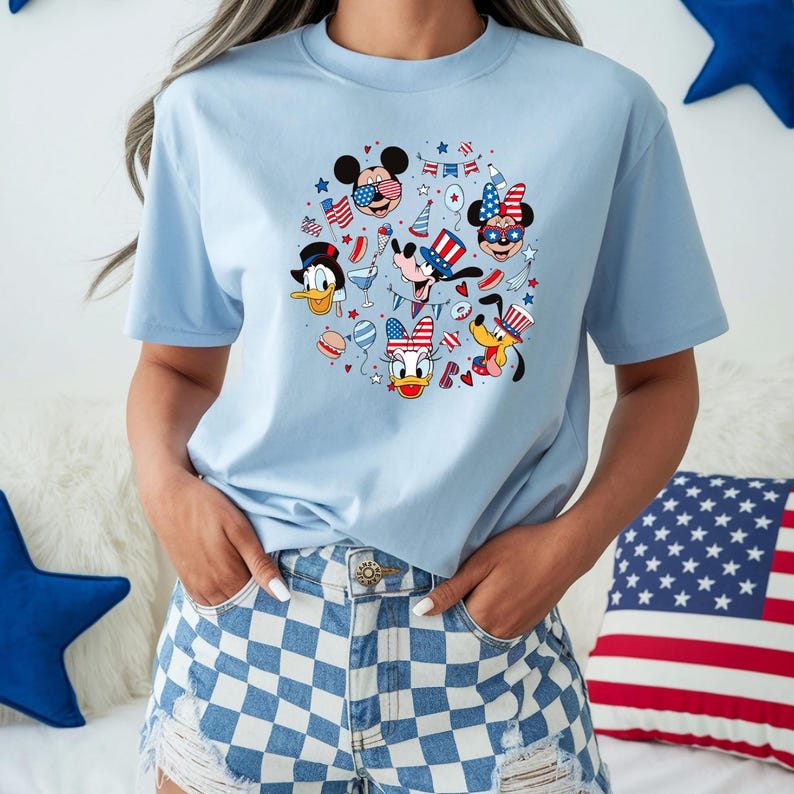May include: Light blue t-shirt featuring a circular graphic of cartoon characters in patriotic attire. The graphic includes Mickey Mouse, Minnie Mouse, Donald Duck, Daisy Duck, Goofy, and Pluto, with American flag-themed accessories. The shirt is paired with blue and white checkered shorts.