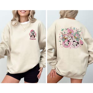 May include: Beige sweatshirt featuring a festive design. The front displays a cartoon character with the word "Custom" below. The back showcases Mickey and Minnie Mouse, Donald and Daisy Duck, and Goofy in Christmas ornaments, surrounded by lights and a small Christmas tree.