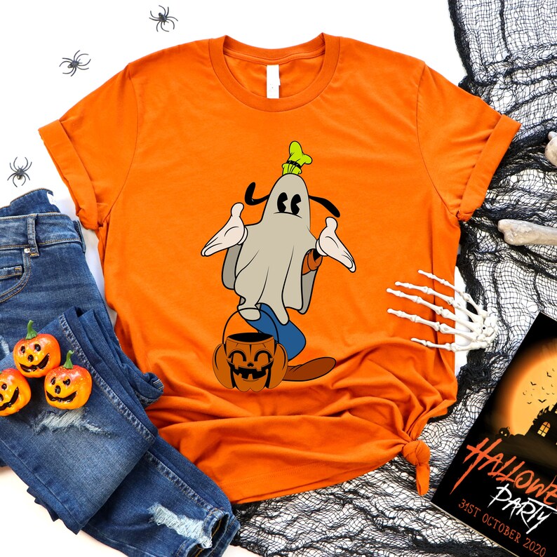 Pode incluir: Camiseta laranja com um personagem de desenho animado vestido de fantasma segurando um balde de ab&oacute;bora. A imagem inclui jeans azuis, ab&oacute;boras em miniatura, uma m&atilde;o de esqueleto e um p&ocirc;ster de festa de Halloween.