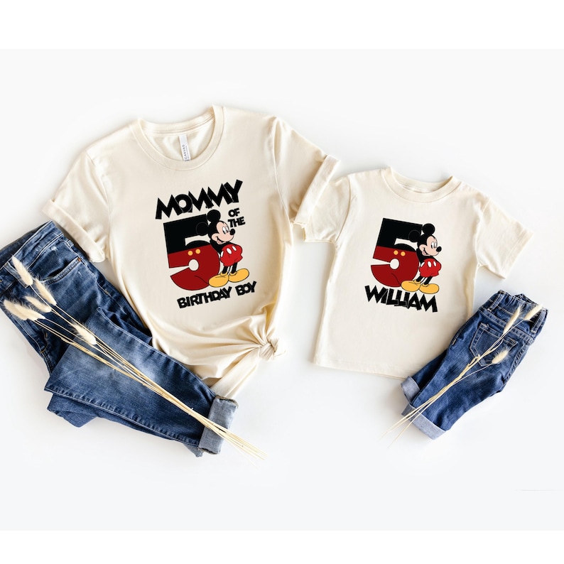 Puede incluir: Dos camisetas color crema con gr&aacute;ficos de Mickey Mouse. Una camiseta dice "Mommy of the Birthday Boy" y la otra "William". Ambas camisetas presentan un gran n&uacute;mero 5 y Mickey Mouse. Vaqueros azules est&aacute;n al lado de las camisas.