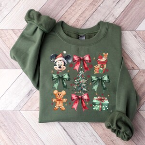 May include: A green sweatshirt with a Christmas theme featuring Mickey Mouse, a Christmas tree, presents, and gingerbread men. The sweatshirt has a crew neck and long sleeves.
