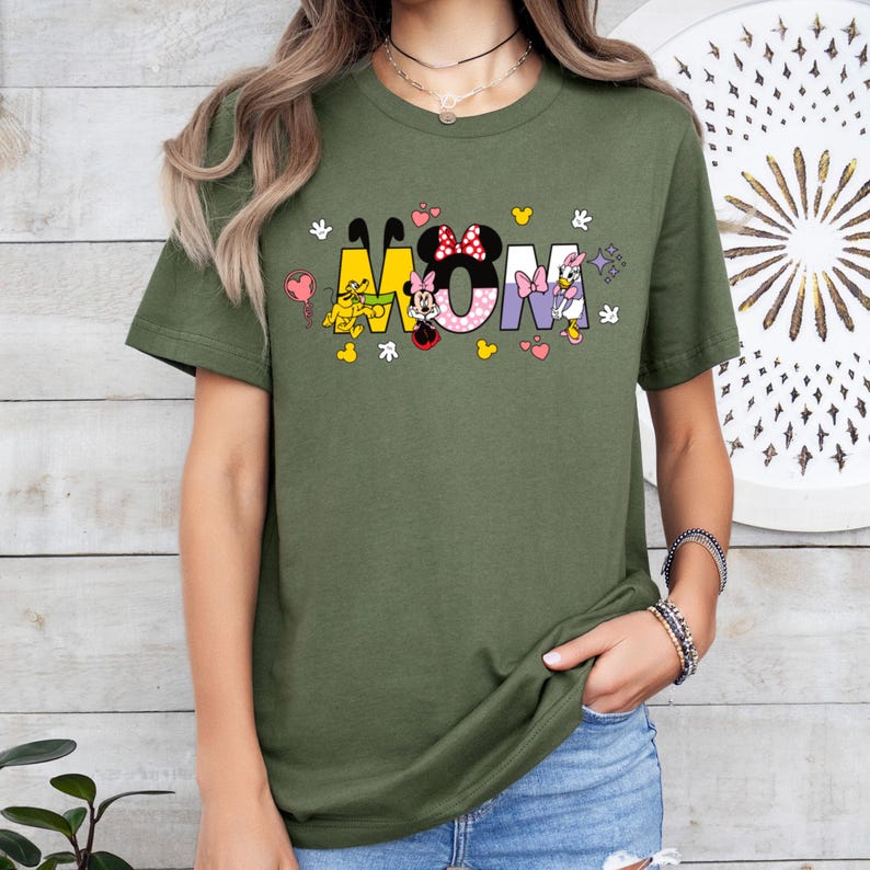 May include: Green short-sleeved t-shirt with a graphic of cartoon characters spelling out "MOM". The characters are Mickey Mouse, Minnie Mouse, Donald Duck, and Pluto.