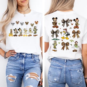 May include: White t-shirt with a print of Disney characters dressed in safari outfits. The front of the shirt features the characters holding balloons with animal print designs. The back of the shirt features the characters with bows and other safari-themed designs.