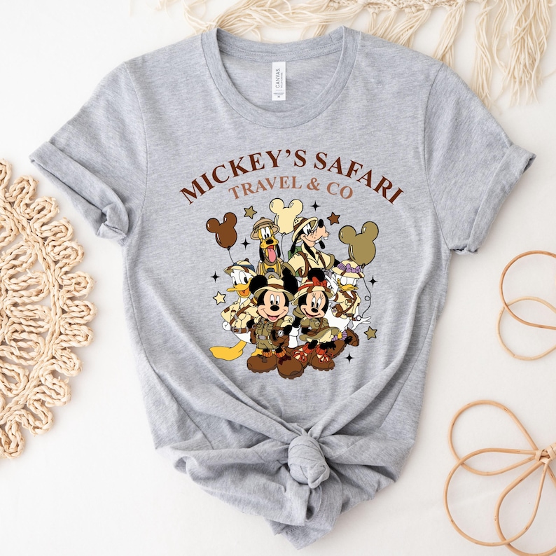 K&ouml;nnte beinhalten: Graues T-Shirt mit einem Aufdruck von Mickey Mouse und seinen Freunden in Safari-Kleidung. Der Text "Mickey's Safari Travel & Co" ist auf dem Shirt gedruckt.