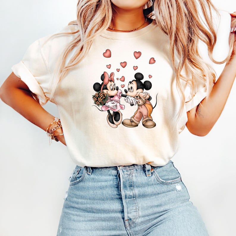 May include: A white t-shirt with a graphic of Mickey Mouse and Minnie Mouse holding hands with hearts floating around them.