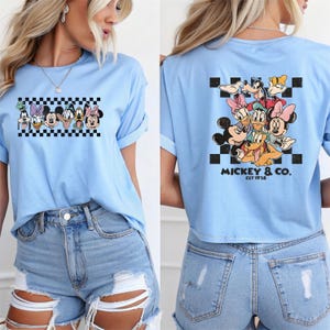 May include: Light blue t-shirt featuring a graphic of Disney characters on a checkered background. The back of the shirt displays a similar design with the text "Mickey & Co. EST 1928".