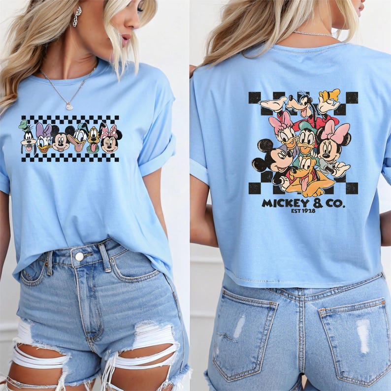 May include: Light blue t-shirt featuring a graphic of Disney characters on a checkered background. The back of the shirt displays a similar design with the text "Mickey & Co. EST 1928".
