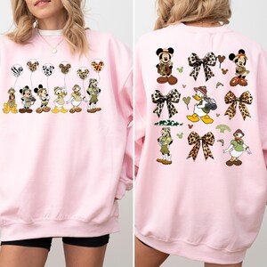 May include: Pink sweatshirt with a graphic print of Mickey Mouse and friends dressed in safari outfits. The print features Mickey Mouse, Minnie Mouse, Donald Duck, Goofy, and Pluto holding balloons with animal print designs. The back of the sweatshirt features a similar design with Mickey Mouse and friends wearing safari hats and holding balloons with animal print designs.