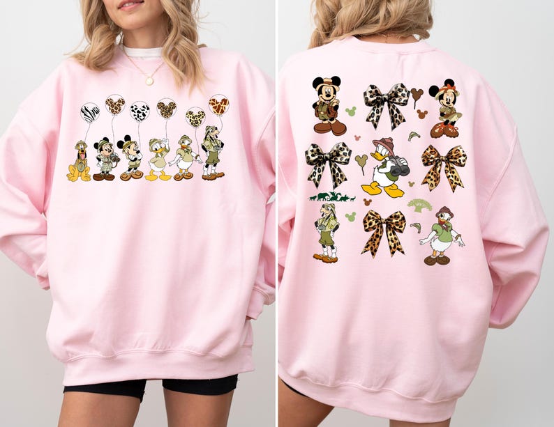 May include: Pink sweatshirt with a graphic print of Mickey Mouse and friends dressed in safari outfits. The print features Mickey Mouse, Minnie Mouse, Donald Duck, Goofy, and Pluto holding balloons with animal print designs. The back of the sweatshirt features a similar design with Mickey Mouse and friends wearing safari hats and holding balloons with animal print designs.