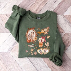 Puede incluir: Sudadera verde con un gr&aacute;fico tem&aacute;tico de oto&ntilde;o con un castillo, Chip y Chop, Mickey y Minnie Mouse y un caf&eacute; con leche de calabaza especiada. El dise&ntilde;o incluye hojas de oto&ntilde;o y el texto "Good Vibes".