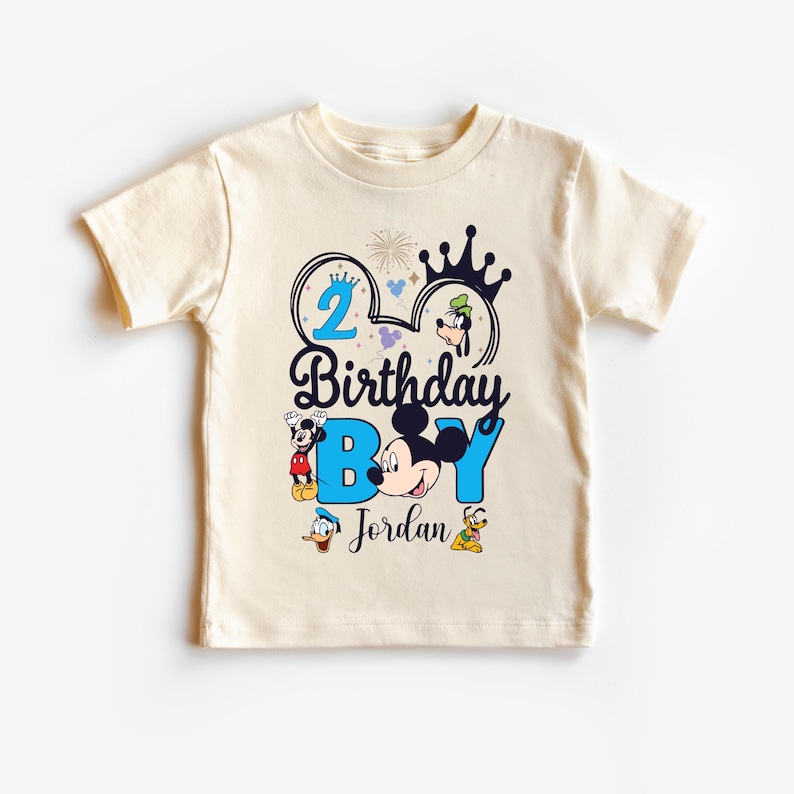 Op de afbeelding: Een cr&egrave;mekleurig T-shirt met een blauw en zwart Mickey Mouse-ontwerp. Het shirt zegt "2 Birthday Boy Jordan" met een kroon en vuurwerk.