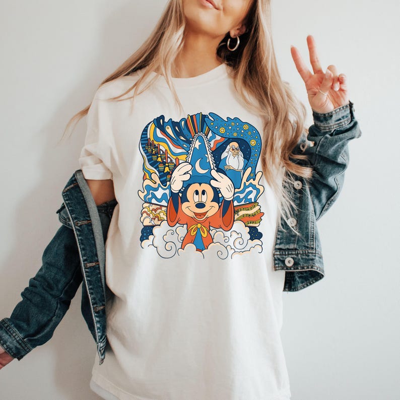 May include: White t-shirt with a colorful graphic of Mickey Mouse as a sorcerer. The graphic includes a blue hat, red robe, and a cloud background. The graphic also includes a castle, fireworks, and a wizard.