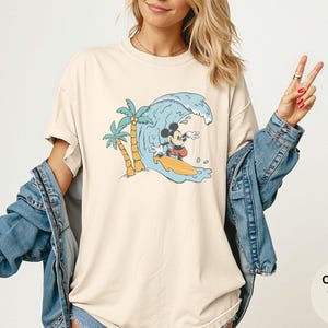 May include: Ivory-colored t-shirt featuring a cartoon Mickey Mouse surfing a wave. The design includes palm trees and a yellow surfboard. The shirt has the words "Comfort Colors Ivory" on the bottom right.